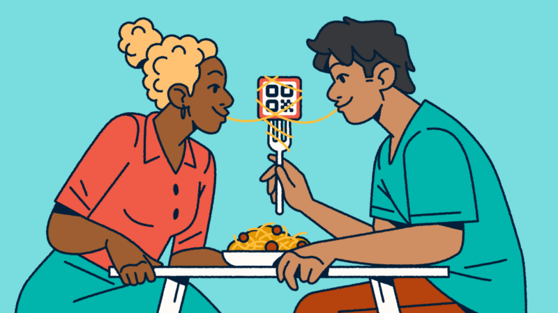 A couple sitting at a table sharing a bowl of spaghetti and meatballs. One partner is holding a fork with a QR Code on it and a piece of spaghetti wrapped around it. They each have one end of the noodle in their mouth.