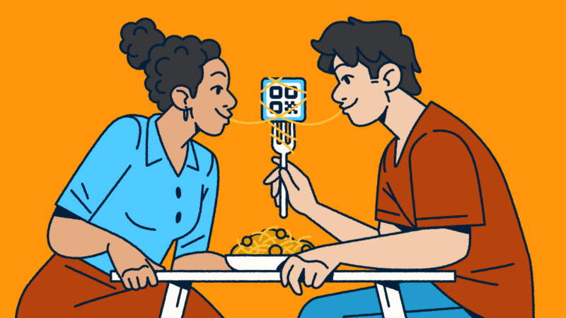 A couple sitting at a table sharing a bowl of spaghetti and meatballs. One partner is holding a fork with a QR Code on it and a piece of spaghetti wrapped around it. They each have one end of the noodle in their mouth.